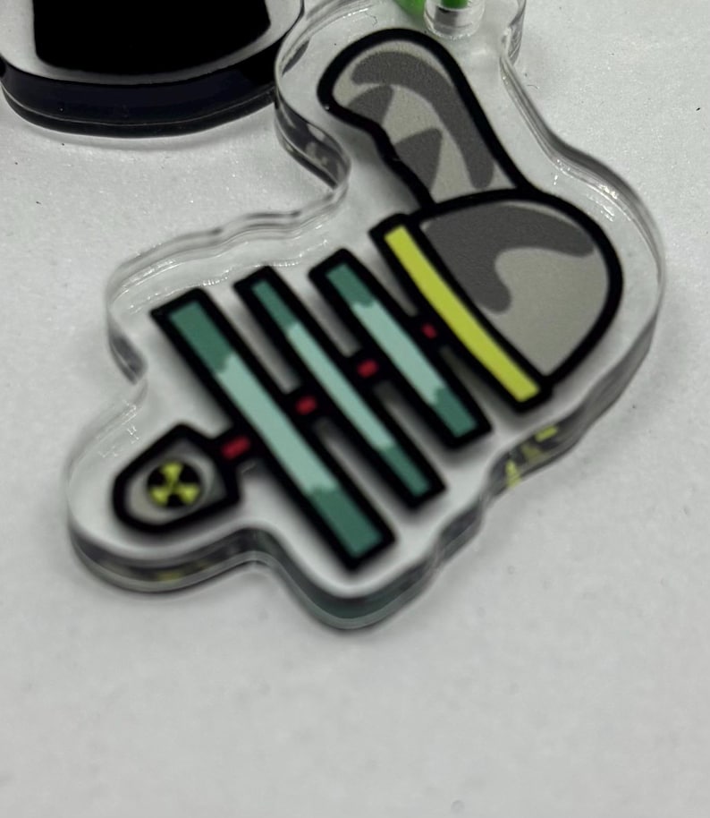 Puede incluir: Un llavero acr&iacute;lico transparente con un dise&ntilde;o de dibujos animados de una espina de pescado y un bolo de bolos. La espina de pescado es de color verde azulado con detalles rojos, y el bolo es gris con detalles amarillos y negros.