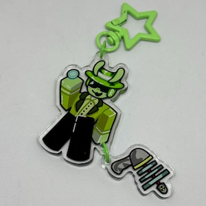 May include: A green and black acrylic keychain featuring a cartoon character in a green suit, hat, and sunglasses, holding a green blaster. The keychain also includes a green star-shaped clip and comes in 2 pieces connected by a jump ring.