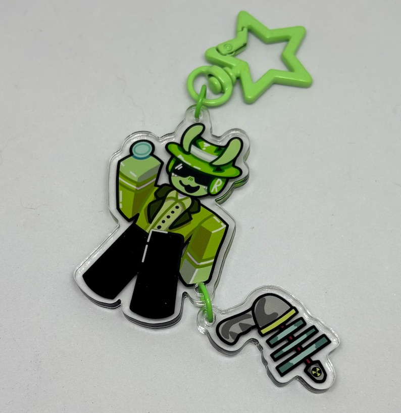 May include: A green and black acrylic keychain featuring a cartoon character in a green suit, hat, and sunglasses, holding a green blaster. The keychain also includes a green star-shaped clip and comes in 2 pieces connected by a jump ring.