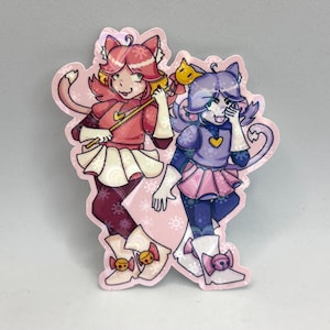 May include: A die-cut sticker featuring two anthropomorphic cat characters. The left character has pink hair, a red top, and a white skirt. The right character has purple hair, a purple top, and a pink skirt. Both wear white boots with bells.