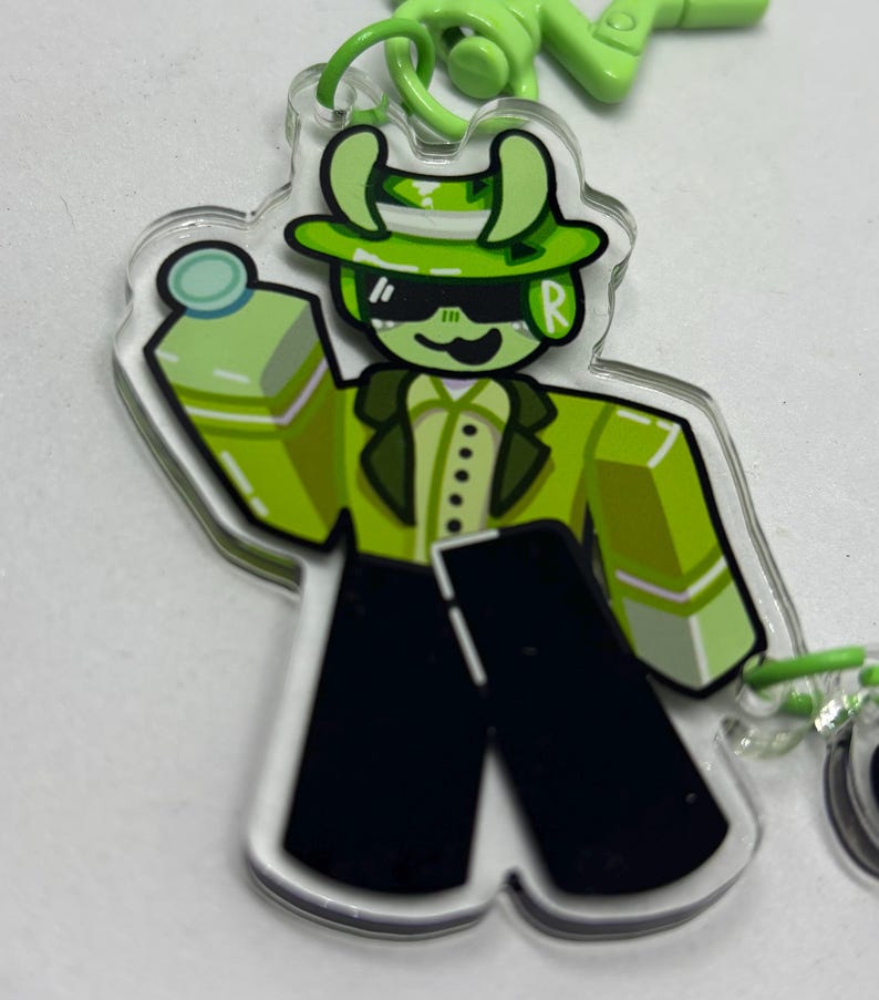 Puede incluir: Un llavero acr&iacute;lico transparente con un personaje de dibujos animados verde con sombrero, gafas de sol y traje. El personaje sostiene una botella verde. El llavero tiene un anillo verde y un cierre verde.