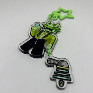 Puede incluir: Un llavero acr&iacute;lico transparente con un personaje de dibujos animados con traje y sombrero verdes, con pantalones negros. El llavero tiene un dije en forma de estrella verde y un segundo dije con un dise&ntilde;o de campana. El personaje sostiene una botella verde.