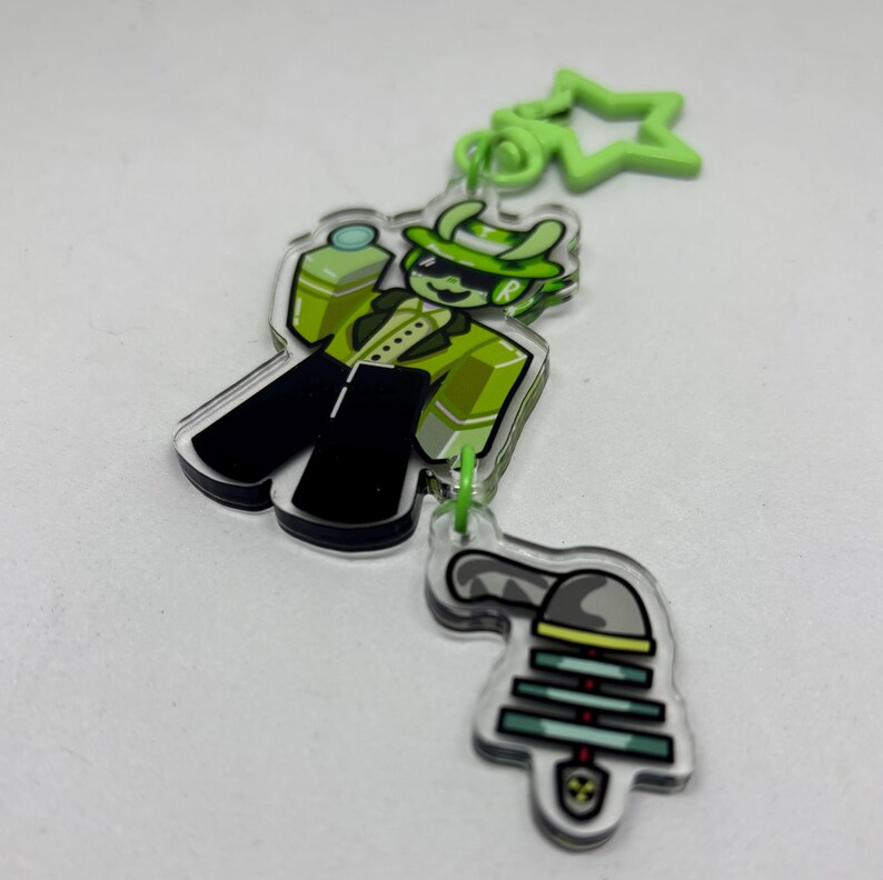 Puede incluir: Un llavero acr&iacute;lico transparente con un personaje de dibujos animados con traje y sombrero verdes, con pantalones negros. El llavero tiene un dije en forma de estrella verde y un segundo dije con un dise&ntilde;o de campana. El personaje sostiene una botella verde.