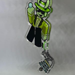 Puede incluir: Un colgante de acr&iacute;lico transparente con un personaje de dibujos animados verde con traje, sombrero y gafas de sol, sosteniendo una peque&ntilde;a bola. El colgante est&aacute; conectado a un colgante m&aacute;s peque&ntilde;o con un lazo verde.