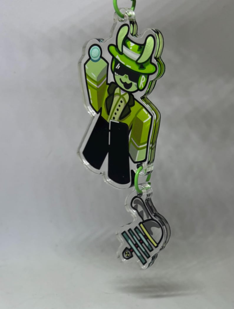 Puede incluir: Un colgante de acr&iacute;lico transparente con un personaje de dibujos animados verde con traje, sombrero y gafas de sol, sosteniendo una peque&ntilde;a bola. El colgante est&aacute; conectado a un colgante m&aacute;s peque&ntilde;o con un lazo verde.