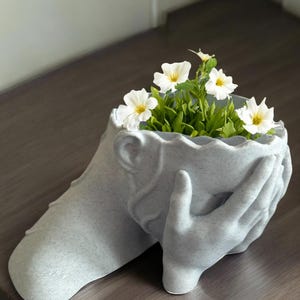 May include: A gray ceramic planter shaped like a person with their hands covering their face. The planter is filled with green plants and white flowers.