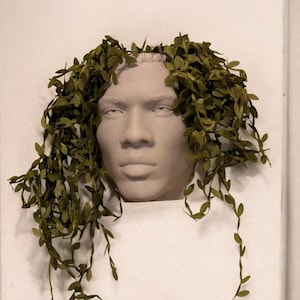 May include: A white plaster sculpture of a person's head with green leafy vines growing from the top, resembling hair.
