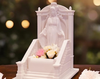 May include: A white religious statue featuring a figure with outstretched arms, set within a decorative architectural frame. The statue has a small planter box filled with pink and white flowers. The base has steps and a rectangular platform.