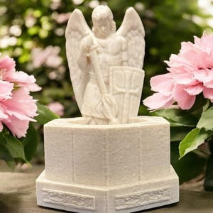May include: A detailed, off-white angel statue with outstretched wings, holding a staff and shield. The statue is set on a textured, octagonal base with decorative trim. Pink flowers and green foliage frame the statue.