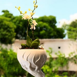 May include: White ceramic planter shaped like a tree trunk with a white orchid plant inside. The planter has a wide base and a narrow top, with the trunk twisting and turning to create a unique design.