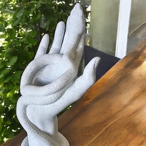 May include: A white ceramic sculpture of a hand holding a snake. The snake is coiled around the hand and its tail is curled up at the base of the hand.