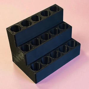 May include: A black, three-tiered display stand with multiple circular openings. The stand is made of a solid material and is designed to hold small items. The background is a solid pink color.