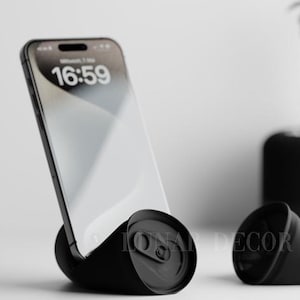 May include: A black phone stand made from a crushed can, holding a smartphone displaying the time 16:59. Another crushed can is next to it. A small potted plant is in the background.