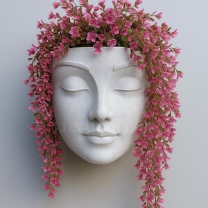 May include: A white, wall-mounted planter shaped like a serene face with closed eyes. The planter is filled with cascading pink flowers, creating a unique and artistic display. Ideal for adding a touch of elegance to any space.