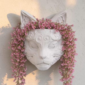 May include: A white, cat-shaped wall planter with intricate facial details and large ears. The planter is filled with cascading pink flowers, creating a whimsical and decorative piece. The planter is mounted on a textured white wall.