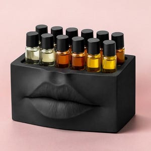 May include: A black rectangular perfume bottle holder with a sculpted lip design. The holder contains several small glass bottles with black caps, filled with various colored liquids. The background is a solid light pink.