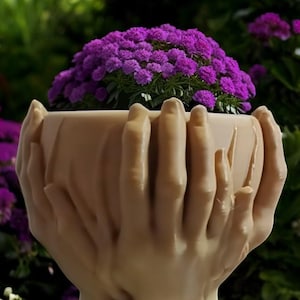 May include: A planter shaped like two hands holding a pot, with a purple flower inside. The hands are made of a light brown material and have a textured surface. The planter is sitting on a wooden surface.