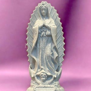 May include: A grey statue of the Virgin Mary, hands clasped in prayer, against a pink backdrop. The statue has a halo and a cherub at the base. A religious item, the statue is a representation of faith and devotion.