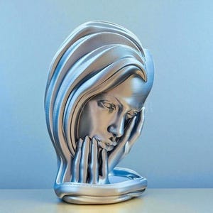 May include: A silver-colored sculpture of a woman with her head resting on her hands. The sculpture has flowing hair and a contemplative expression. The base is a rounded shape. The background is a gradient of blue.