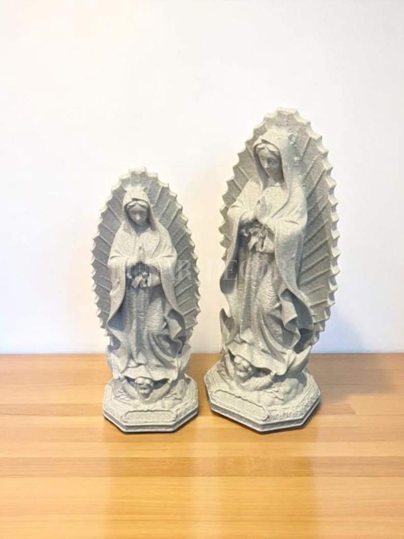 May include: Two grey statues of the Virgin Mary. The statues have detailed flowing robes and a halo. The statues are on a wooden surface.