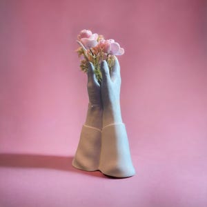 May include: A white ceramic vase shaped like two hands holding a bouquet of pink flowers.