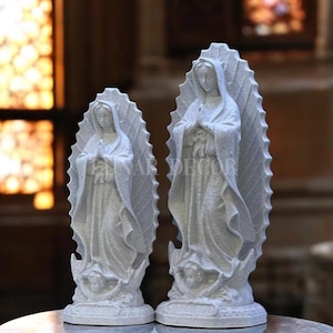 May include: Two white statues of a religious figure, each with a halo-like backdrop. The statues are standing on a round base, with the text "INAR DECOR" visible. The statues are on a marble-like surface.