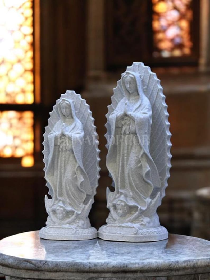 May include: Two white statues of a religious figure, each with a halo-like backdrop. The statues are standing on a round base, with the text "INAR DECOR" visible. The statues are on a marble-like surface.