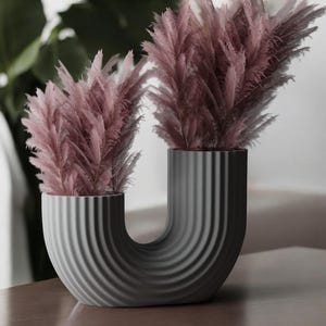 May include: A gray ceramic vase with a unique U-shaped design and vertical ridges. The vase is filled with two bunches of dried pink pampas grass.