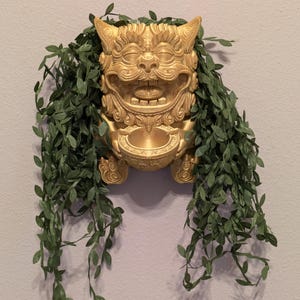 May include: A gold-colored, decorative lion statue with intricate details. The statue is holding a small bowl in its paws. The statue is surrounded by green, leafy vines.