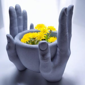 May include: A gray ceramic planter shaped like two hands holding a bowl. The bowl is filled with yellow dandelion flowers.