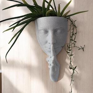 May include: A gray concrete planter shaped like a face with its finger to its lips, as if to say 'shhh'. The planter is filled with green plants.