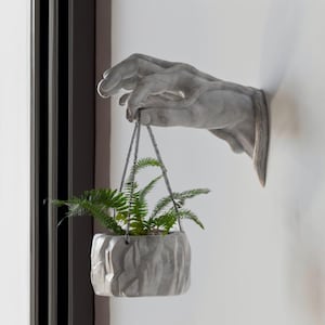 May include: A concrete hand sculpture with a hanging planter. The planter is made of a textured, gray material and contains a green plant.