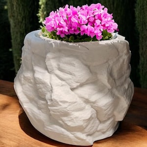 May include: A white ceramic planter shaped like a rock with pink flowers inside.