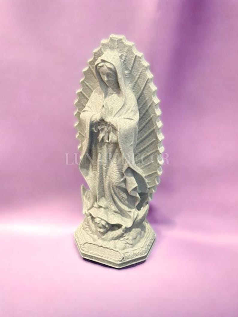 May include: A grey statue of the Virgin Mary, standing with hands clasped in prayer. The statue features a detailed depiction of her robes and a halo-like backdrop. The base is hexagonal with ornate carvings. The background is a solid light purple.