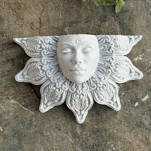 May include: A gray, decorative wall hanging featuring a serene face with closed eyes, surrounded by a detailed, petal-like design. The sculpture has an intricate, carved appearance, suggesting a spiritual or artistic theme. The piece is made of a light gray material.