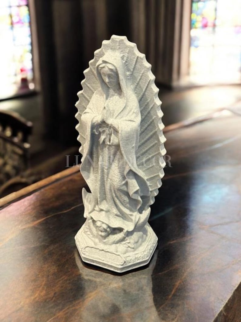 May include: A grey statue of a religious figure, with a halo-like design behind the head. The figure is wearing a draped garment and is standing on a decorative base. The statue is on a dark, polished surface.