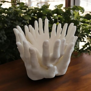 May include: White ceramic bowl shaped like a group of hands cupped together. The bowl is on a wooden surface.