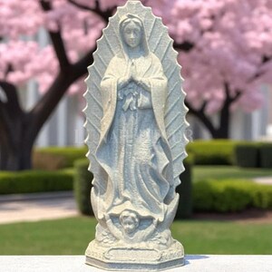 May include: A light grey statue of the Virgin Mary, hands clasped in prayer. The statue has a halo and is set on a base with an angel. The background features a pink flowering tree and green grass.