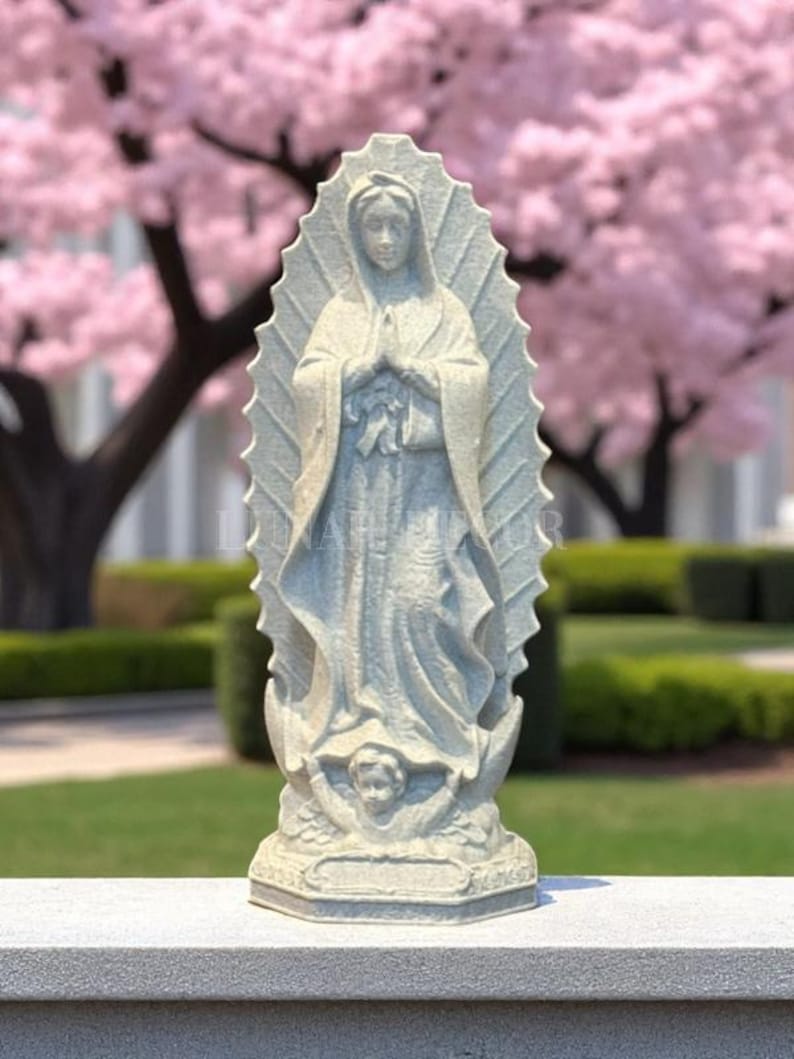 May include: A light grey statue of the Virgin Mary, hands clasped in prayer. The statue has a halo and is set on a base with an angel. The background features a pink flowering tree and green grass.