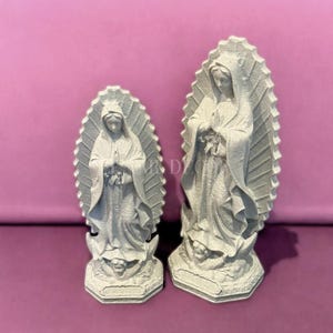 May include: Two gray Virgin Mary statues. The statues are standing, with hands clasped in prayer. Each statue has a halo-like design behind the head. The statues are on a pink background.