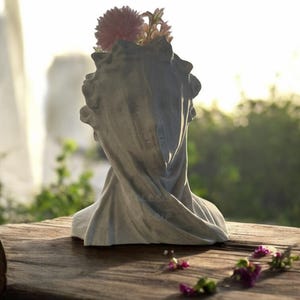 May include: A white plaster bust planter with a draped fabric design. The planter is filled with pink and white flowers.