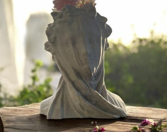 May include: A white plaster bust planter with a draped fabric design. The planter is filled with pink and white flowers.