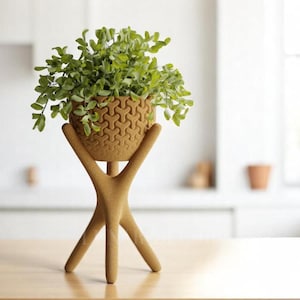 May include: A decorative plant stand with a geometric patterned pot holding a green leafy plant. The stand is made of a light brown material and has three legs. The plant pot is approximately 6 inches in diameter.