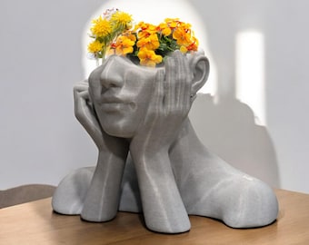 May include: A grey concrete sculpture of a person's head with flowers planted in the top. The sculpture is sitting on a wooden table.