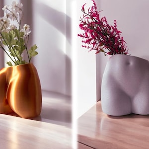 May include: Two ceramic vases shaped like human hips. One vase is gold and holds white flowers. The other vase is gray and holds pink flowers.