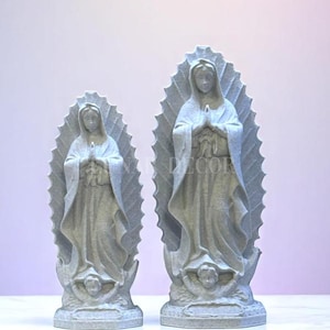 May include: Two grey statues of the Virgin Mary, each with a halo and hands clasped in prayer. The statues are on a white surface, with a light purple background. The statues are of different sizes.