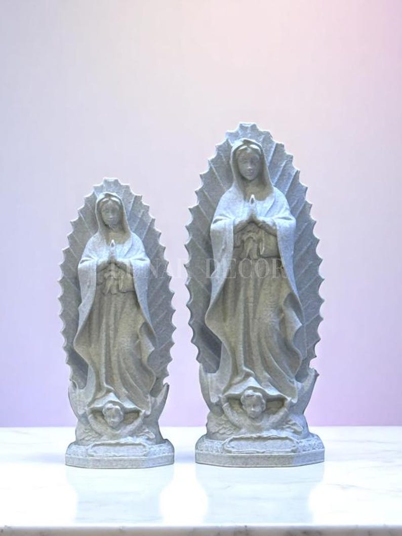 May include: Two grey statues of the Virgin Mary, each with a halo and hands clasped in prayer. The statues are on a white surface, with a light purple background. The statues are of different sizes.