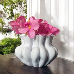 May include: A white ceramic vase with a unique, textured, and organic shape, resembling multiple figures. The vase is filled with pink orchid flowers.
