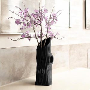May include: A black ceramic vase shaped like a tree trunk with a rough texture, holding a bouquet of pink flowers.