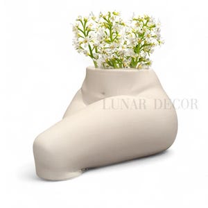 May include: A white ceramic vase shaped like a human torso, with a bouquet of white flowers in the opening. The vase is sitting on a white surface.  The text "LUNAR DECOR" is visible on the vase.